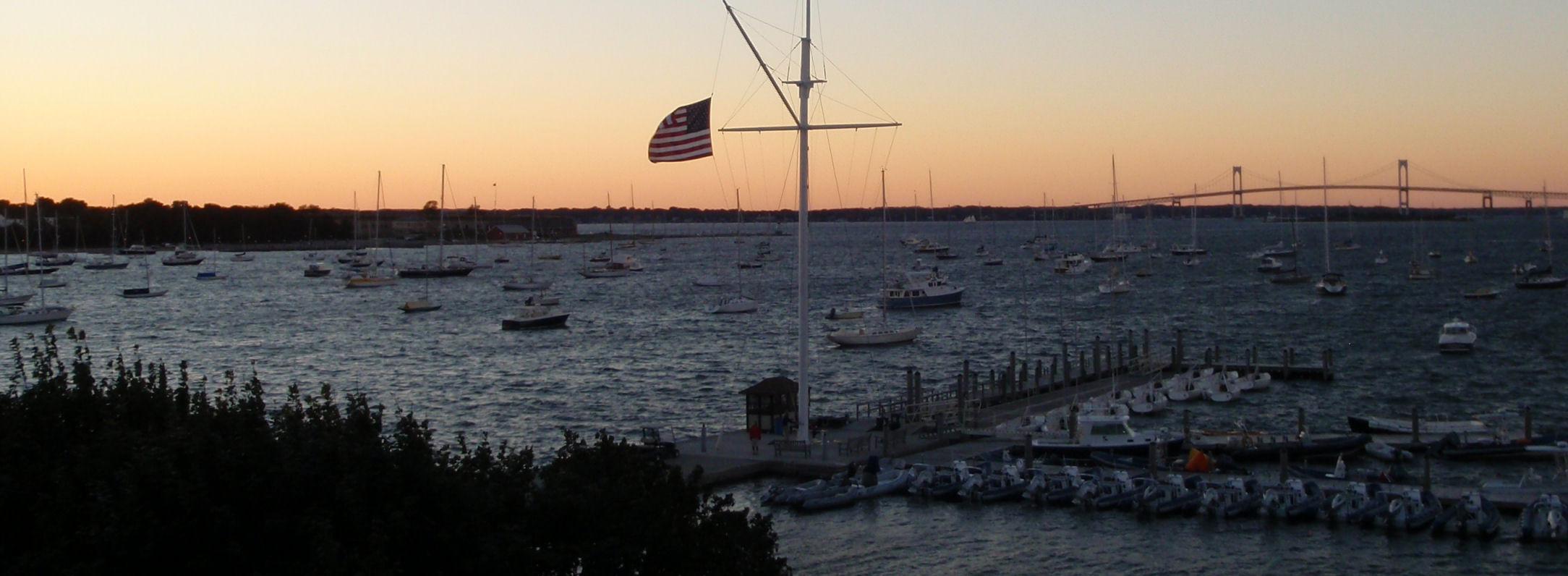 Billedet viser aften solen fra New York Yacht Clubs klubhus her i Newport. De fleste sejladser kommer til at foregå ude ved broen, som anes i baggrunden.